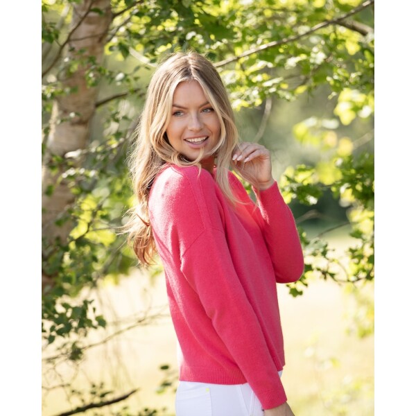 Women’s vibrant coral Teaberry jumper worn with white trousers in a woodland setting